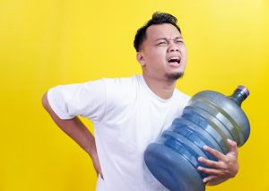 A man struggling to carry a heavy water gallon, holding his back in pain, expressing discomfort.