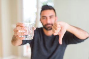  man drinking a fresh glass of water with angry face, negative sign showing dislike with thumbs down, rejection concept. drinking water problems at office concept.
